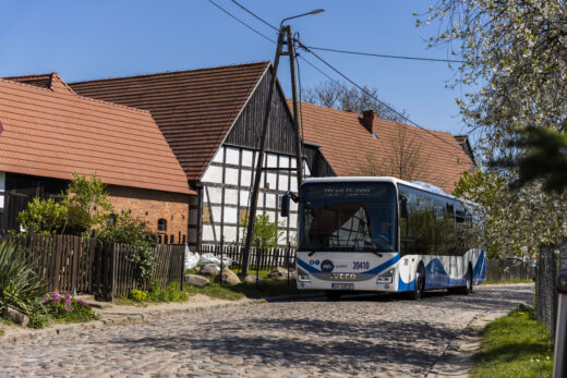Niskopodłogowy autobus PKS Słupsk w zabytkowej wsi Swołowo wśród szachulcowej zabudowy i kwitnących drzewek, poruszający się drogą wyłożoną kocimi łbami.